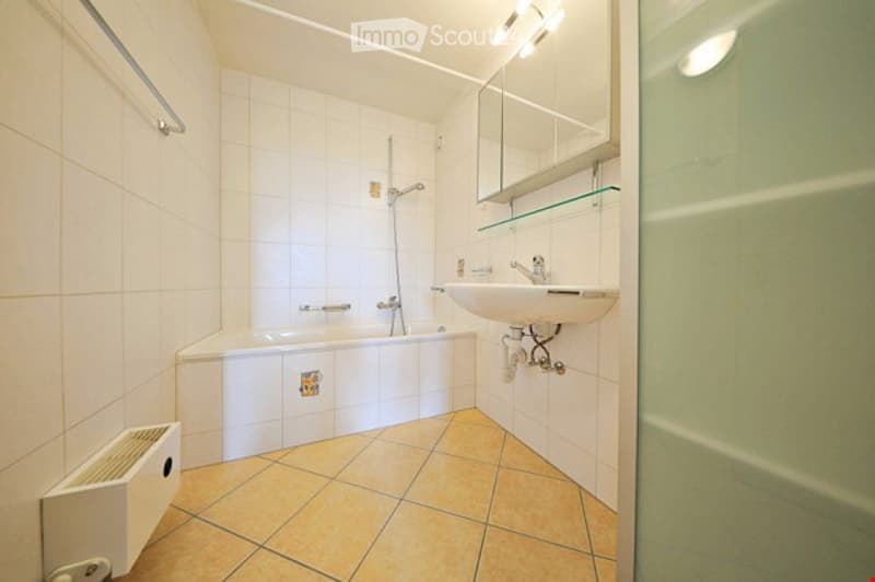 Modern bathroom with white tiled walls, a bathtub, a sink with a mirror, and a radiator. The floor is tiled in a warm beige color.