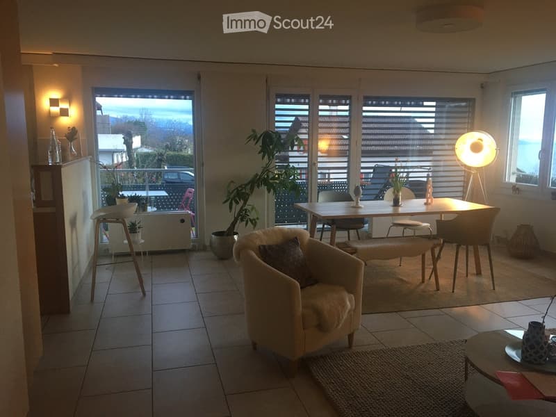 Bright living room with large windows and a view, featuring a cream-colored sofa, coffee table, dining table with chairs, and decorative vases.