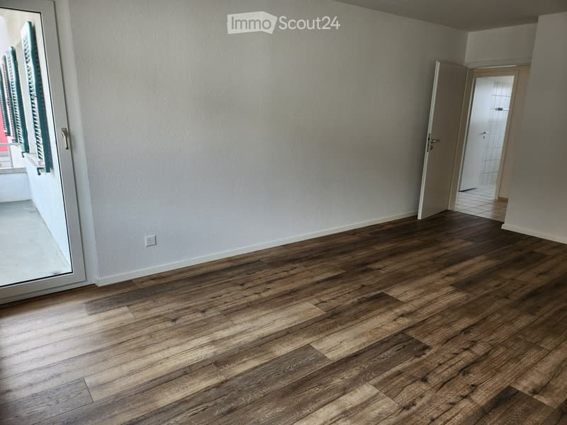 Empty room with wooden flooring, white walls, and a large window with shutters. A doorway leads to another room with tiled flooring.
