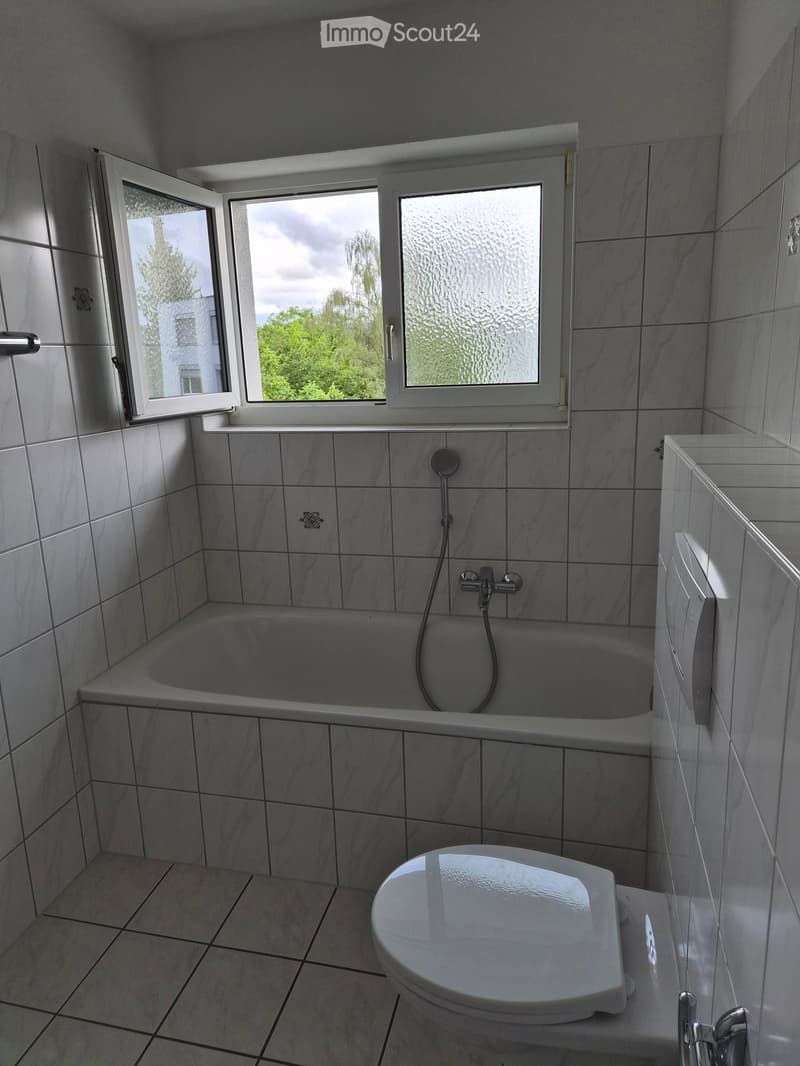 Modern bathroom with white tiled walls and floor, featuring a bathtub, toilet, and a window with frosted glass. The space is clean and well-lit with natural light from the window.