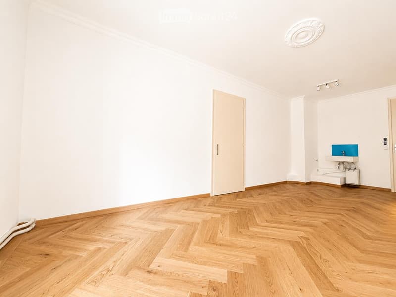 Empty room with light wooden herringbone flooring, white walls, and a small built-in sink area in the corner.