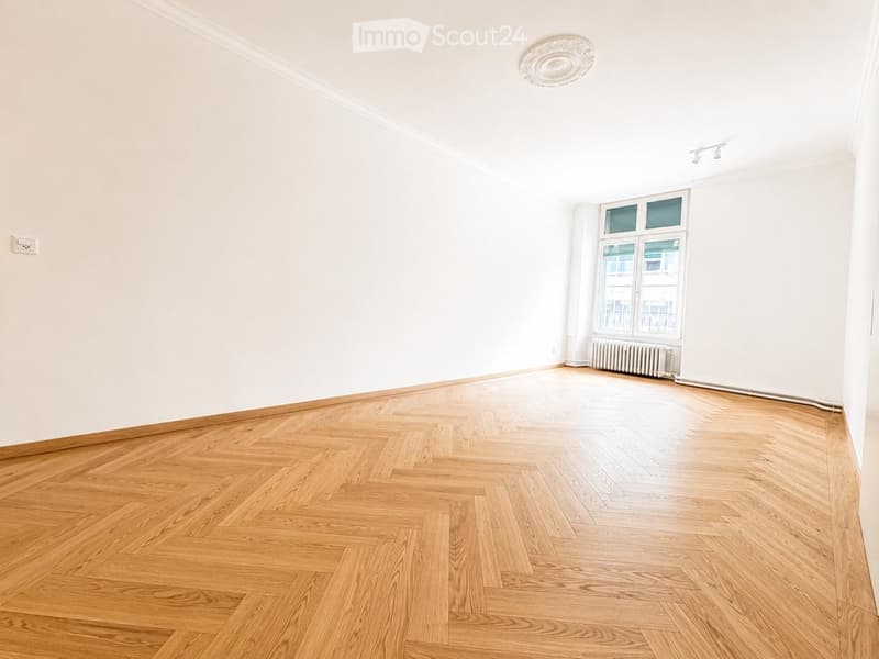 Empty room with light-colored wooden herringbone flooring, white walls, and a large window with a radiator below. The ceiling features a decorative light fixture and track lighting.