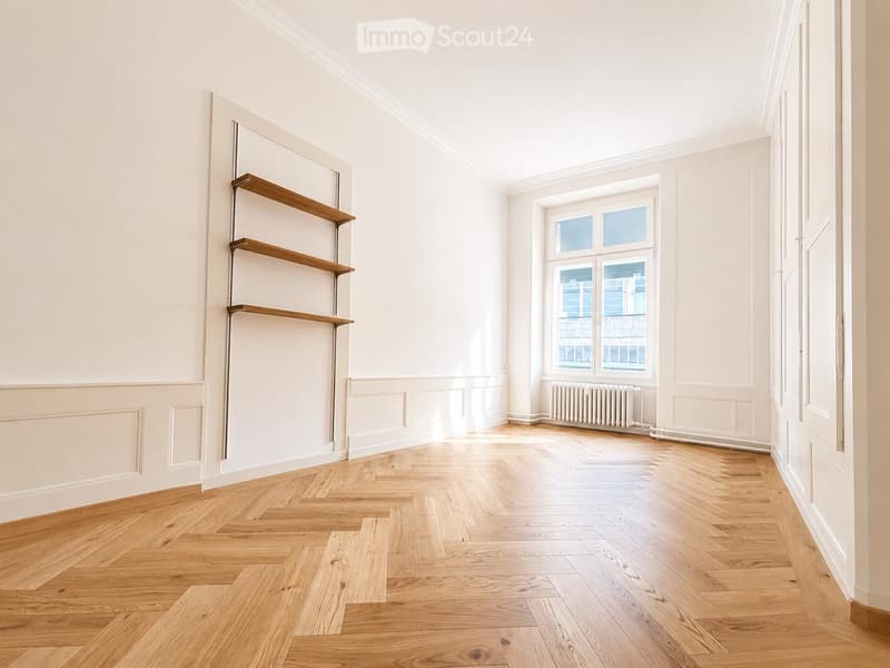 Empty room with light wooden flooring, white walls, and built-in wooden shelves on one wall. A large window with multiple panes allows natural light to enter, and a radiator is visible beneath the window.