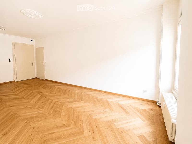 Empty room with light wooden herringbone flooring, white walls, and two doors. Natural light streams in from a window on the right side.