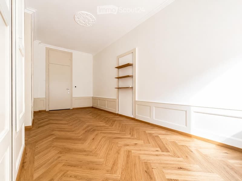 Empty room with light-colored walls, wooden herringbone flooring, and a small built-in shelf unit.