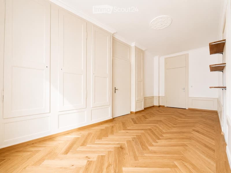 Empty hallway with light wooden herringbone flooring, white walls, and several closed doors.