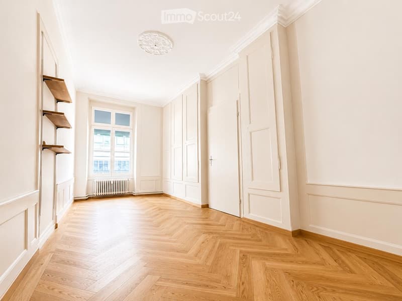 Empty room with light-colored wooden herringbone flooring, white walls, and a large window with a radiator below. The room features built-in cabinetry and a decorative ceiling light fixture.