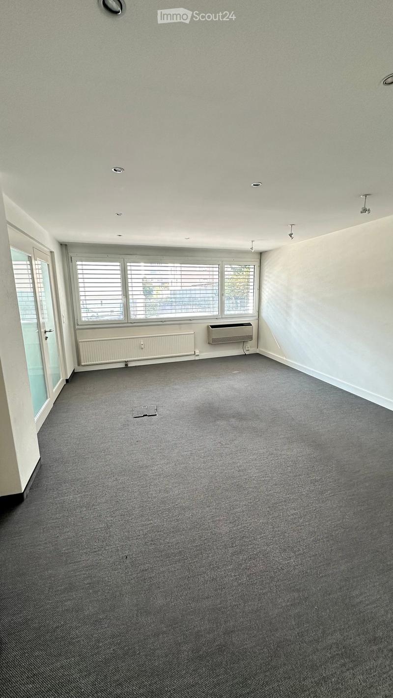 Empty room with grey carpet, white walls, large windows with blinds, and air conditioning units on the wall.