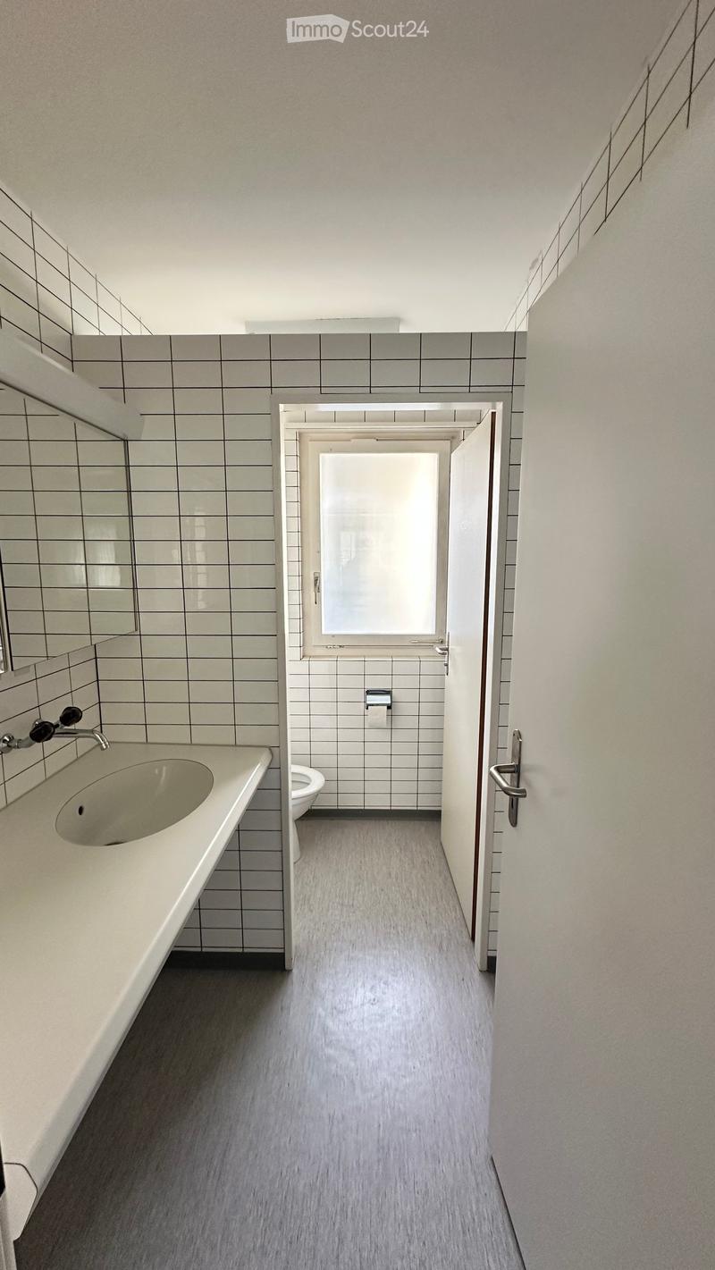 Modern bathroom with a sink, faucet, mirror, toilet, and paper towel holder. The space features grey tiled walls and a grey floor.