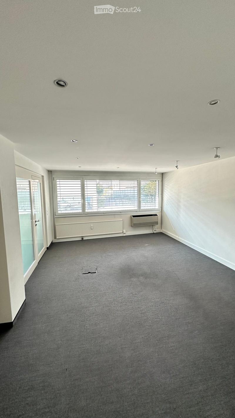 Empty room with grey carpet, white walls, large windows with blinds, and a door leading outside. No furniture or equipment present.
