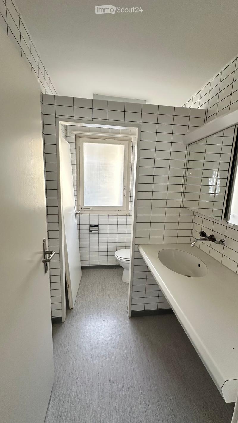 Modern bathroom with grey tiled walls, a white sink with a chrome faucet, and a toilet in an adjacent room.