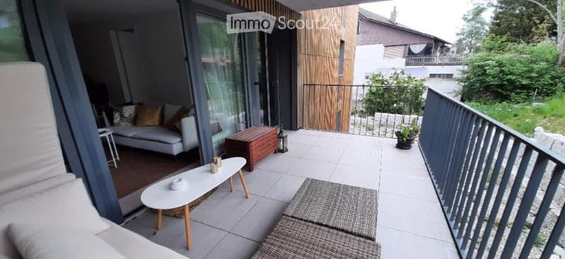 Modern balcony with white sofa, coffee table, wooden storage box, and small potted plant. Large glass sliding doors open to the interior.