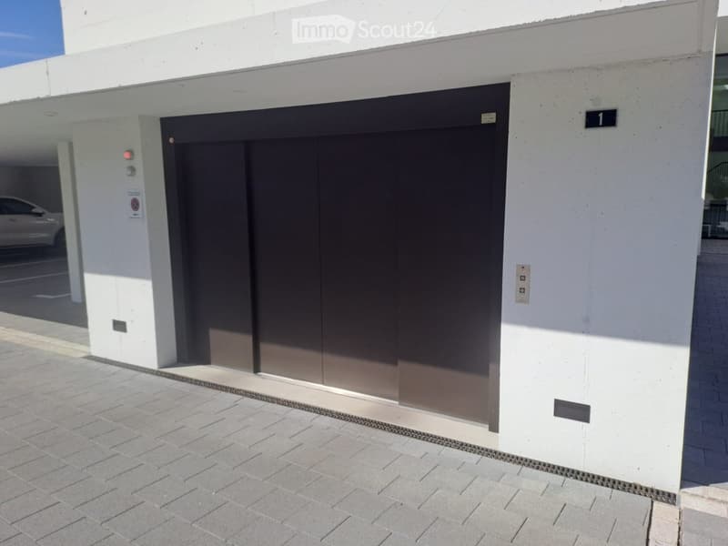 Modern building entrance with a large, dark-colored garage door and a white facade. The entrance features a tiled pavement and a small sign indicating the building number.