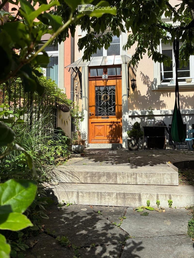 Front entrance of a house with a wooden door, small porch, and surrounding greenery.