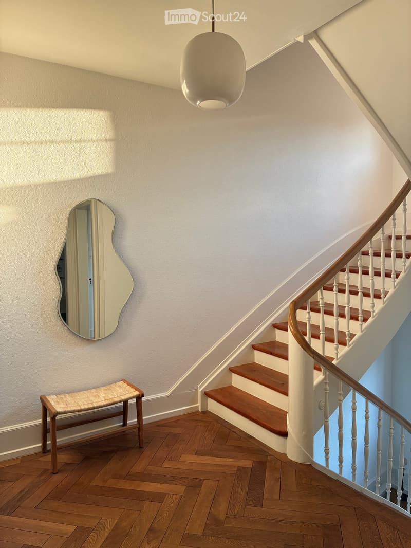 Spacious hallway with wooden flooring, a curved staircase with wooden steps and white railings, a hanging pendant light, a small wooden bench, and a decorative mirror on the wall.