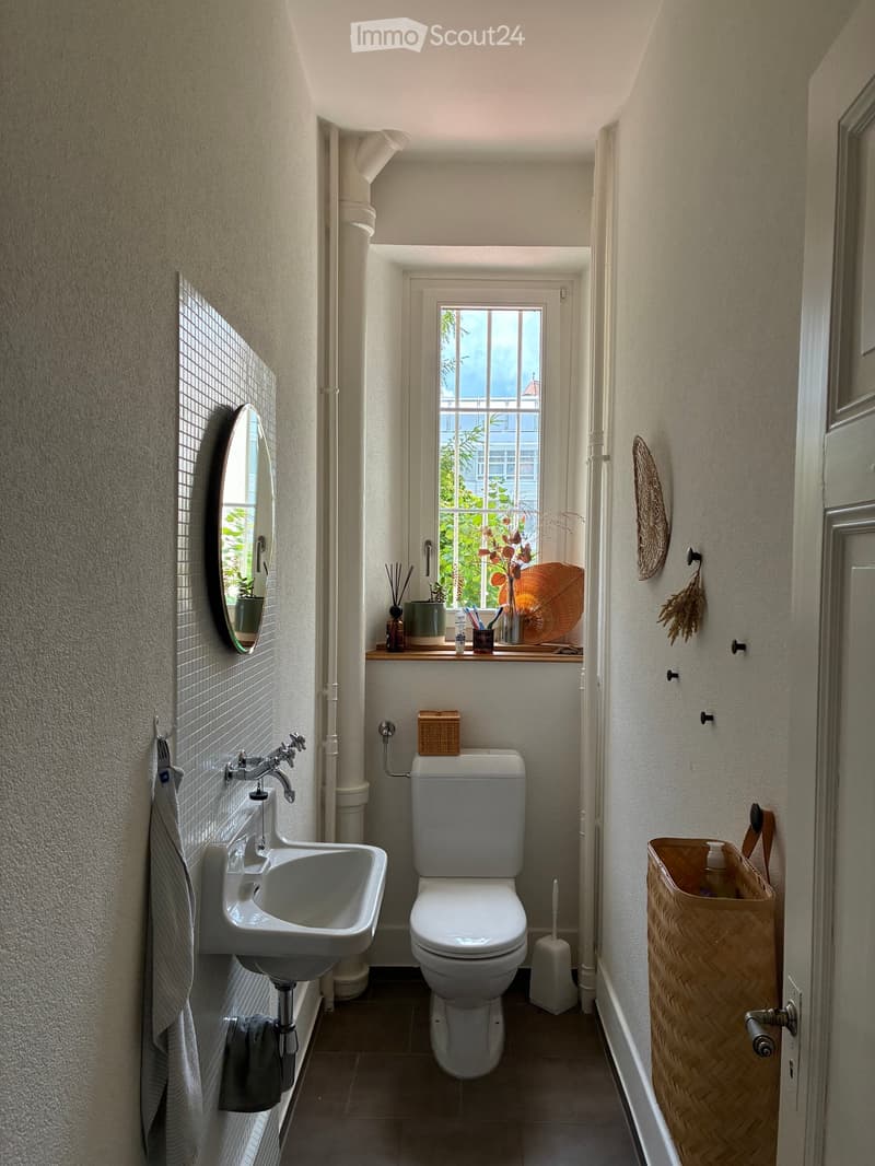 A compact bathroom with a white sink, toilet, and a window offering a view of greenery. The space includes a mirror, a towel, and a small basket on the floor.