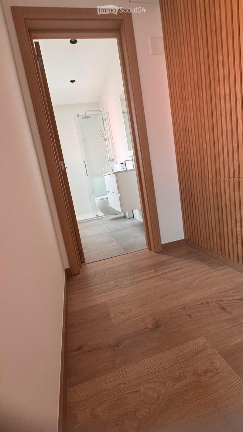 A narrow hallway with wooden flooring and light-colored walls, leading to a modern bathroom with a glass shower enclosure, toilet, and vanity.