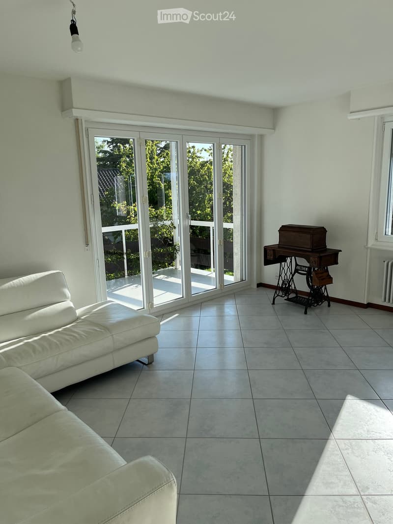 Bright living room with white walls, tiled floor, large sliding glass doors leading to a balcony, a white leather sofa, a small wooden table with a stool, and a hanging light fixture.