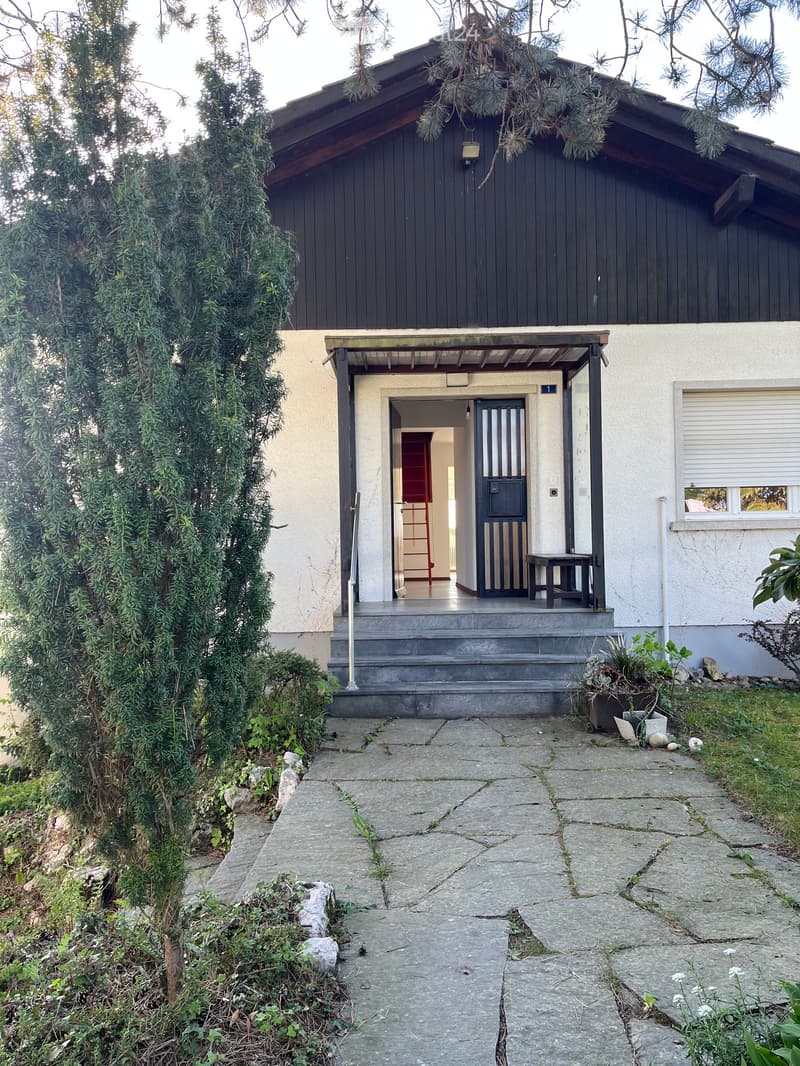 Two-story house with dark wooden exterior, white siding on lower level, covered entryway, small porch with a table, and steps leading up to the door.