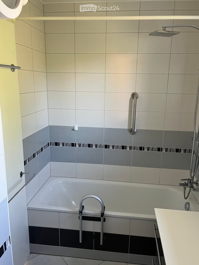 Modern bathroom with a bathtub, showerhead, and handrail. The walls are tiled in a combination of light beige and grey tiles, with a black accent border. A small white countertop is visible on the right side.