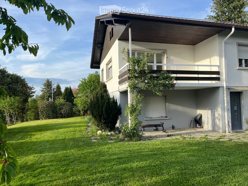 Two-story house with a modern design, featuring a balcony on the upper level, surrounded by a well-maintained lawn and trees.