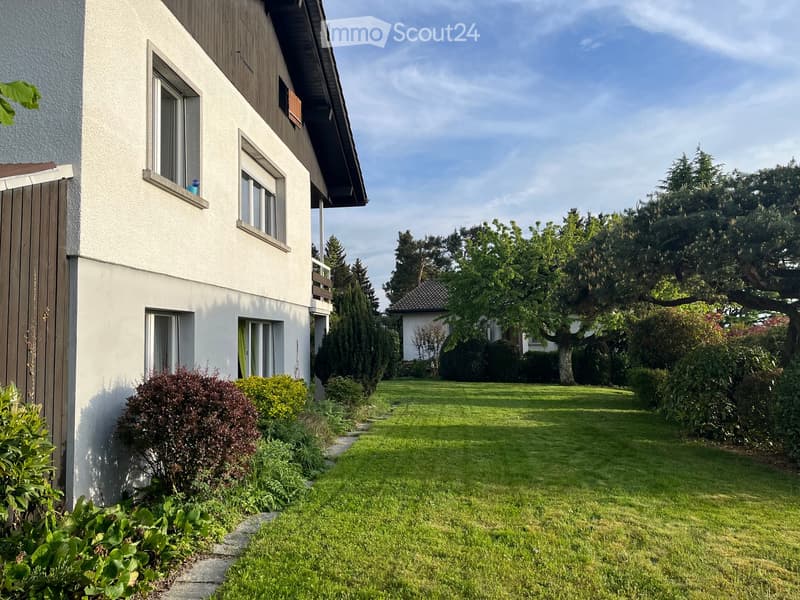 Well-maintained garden with a lawn, shrubs, and trees, adjacent to a two-story house with white walls and wooden accents.
