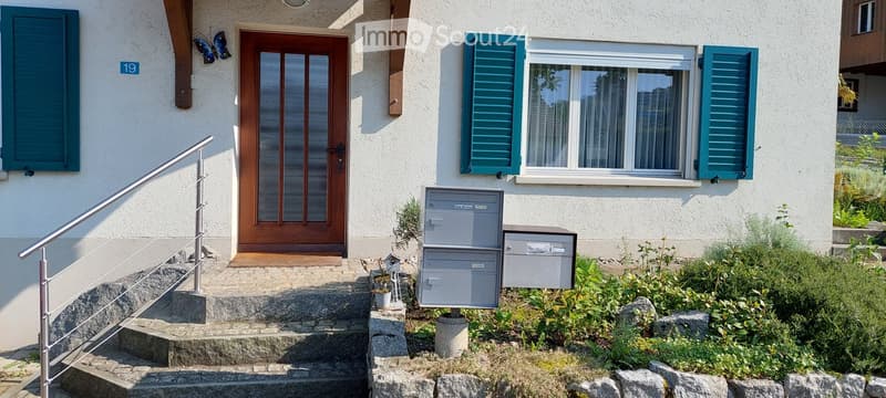 Two-story house with a raised ground floor, featuring a wooden front door, green shutters, and a small garden area with mailboxes.