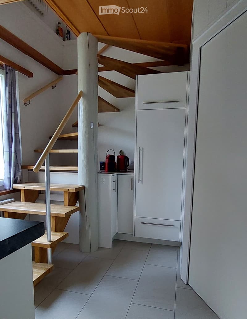 A modern, well-lit hallway with a staircase leading upwards, featuring white tiled flooring, a wooden staircase with a metal railing, and a small kitchen area with white cabinets and a refrigerator.