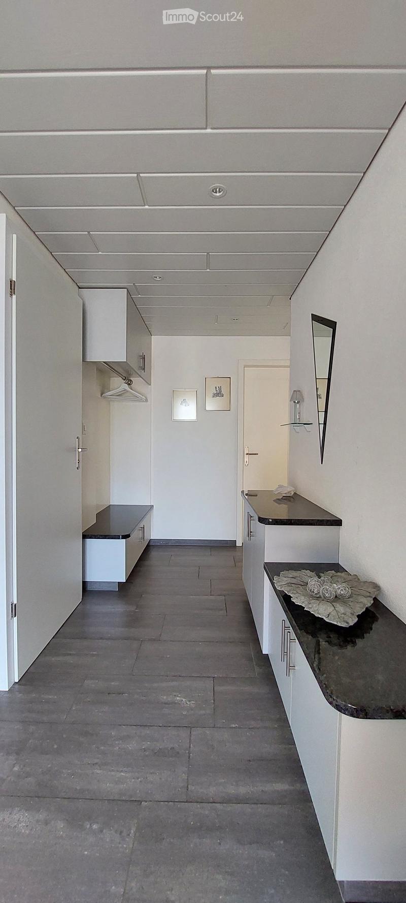 A modern, minimalist hallway with white walls, grey tiled floor, and built-in storage units on both sides. Features include a coat rack, framed artwork, and a small bench.