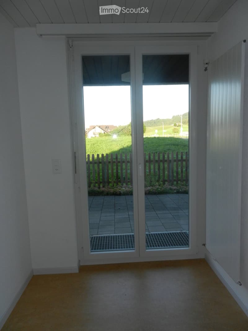 Empty room with light-colored walls and wooden flooring, featuring large sliding glass doors that open to a tiled patio and a view of a grassy area with a wooden fence.