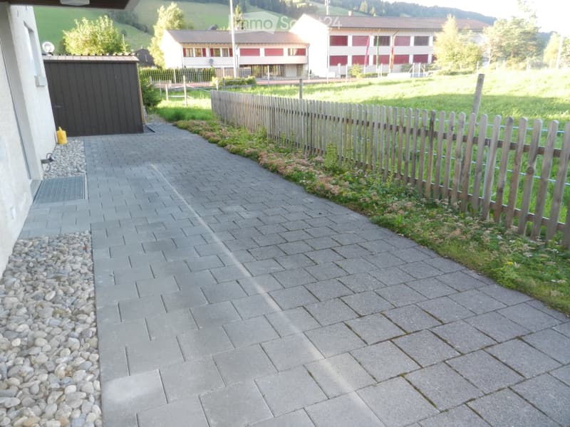 A paved pathway bordered by a wooden fence and small garden area, with a building and distant landscape visible.