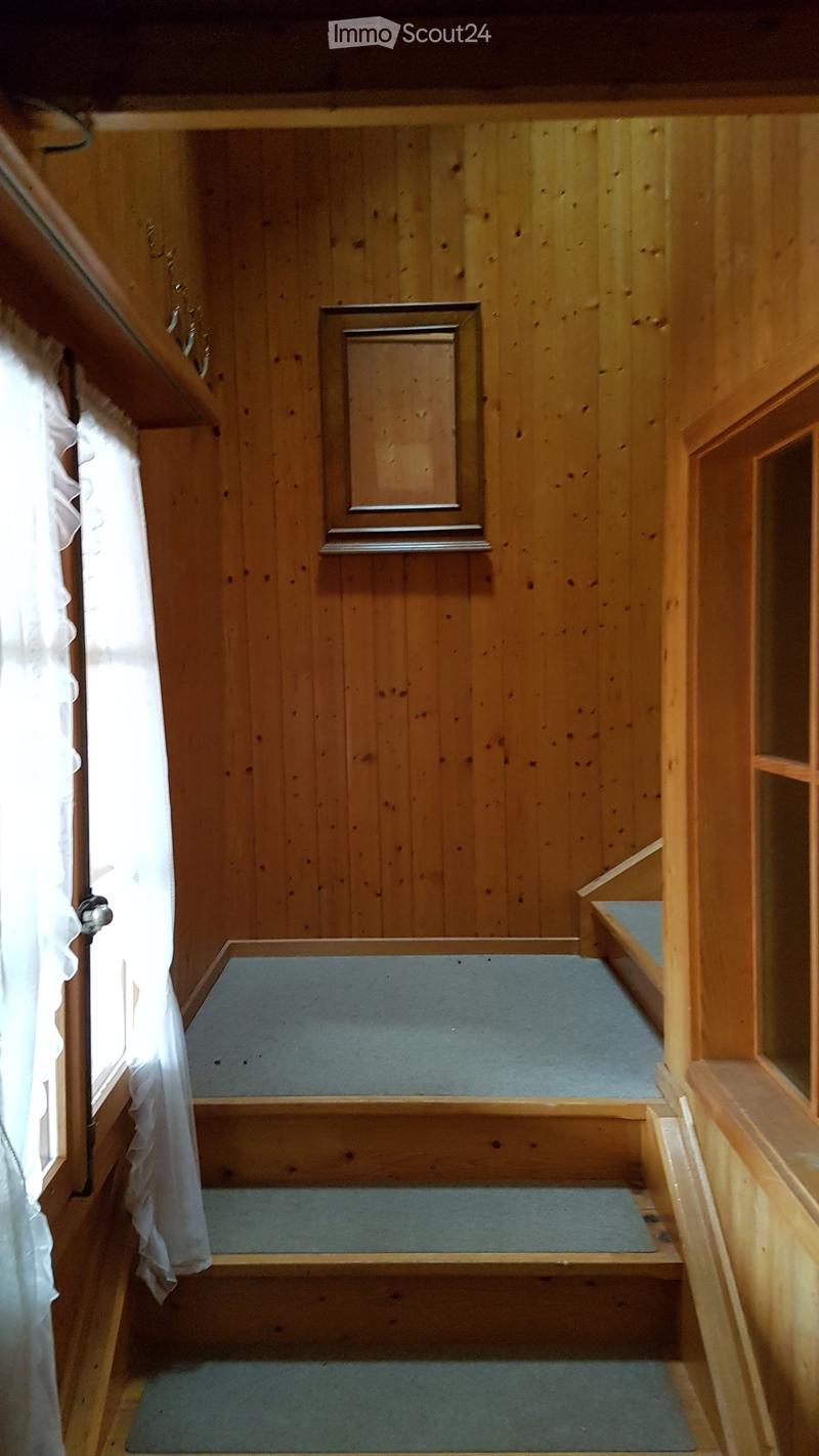 Wooden-paneled hallway with a raised ground floor, featuring a carpeted staircase leading upwards, a window with white curtains, and a small wooden door.