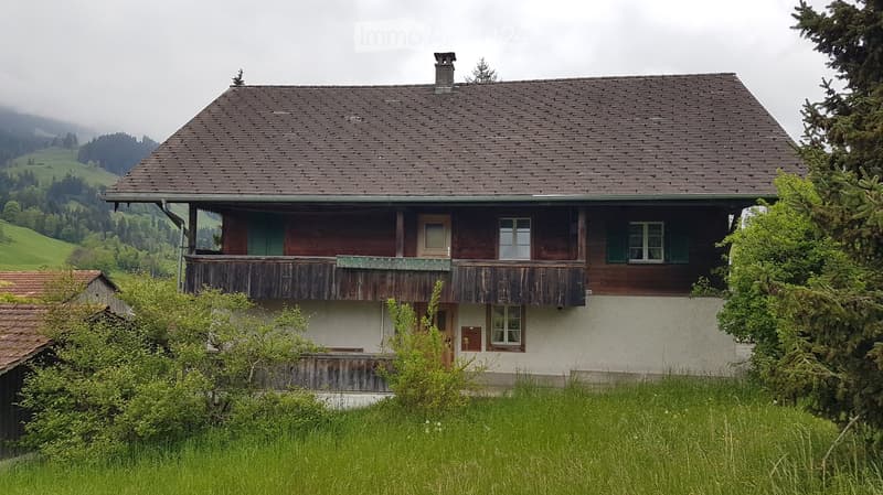 Traditional two-story wooden house with a sloped roof, green shutters, and a balcony overlooking a grassy field and forested hills.