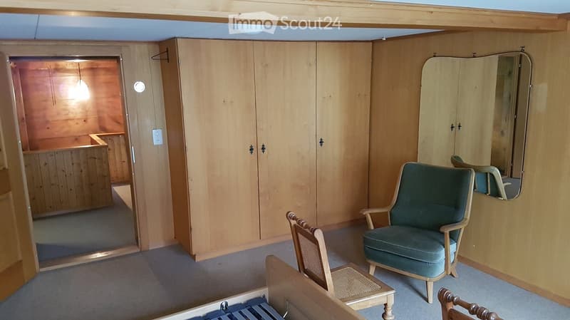 A small, furnished room with wooden walls and a grey carpet. It features a green upholstered armchair, a small wooden table, a large mirror, and a built-in wardrobe with double doors.