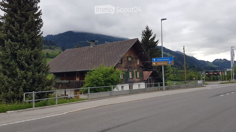 Traditional wooden house with green shutters and a sloped roof, situated near a road with a signpost and surrounding trees and mountains.