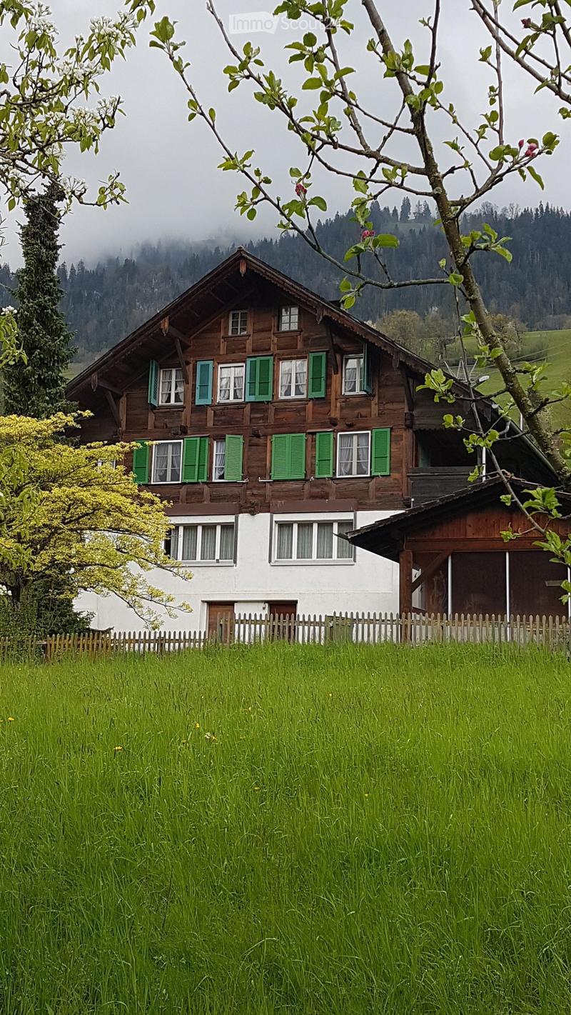 Traditional wooden house with green shutters and a pitched roof, surrounded by a grassy field and trees.