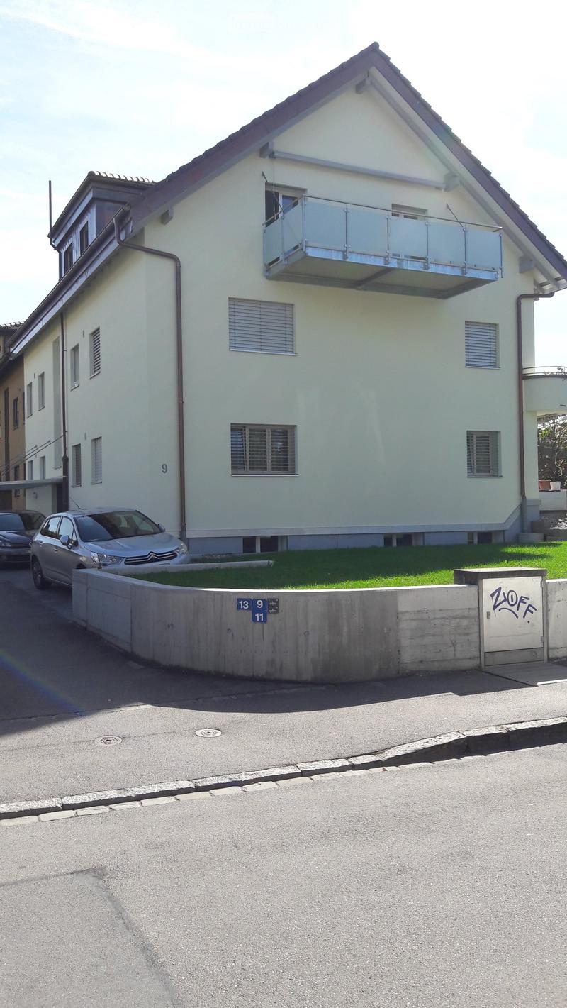 Two-story house with light-colored exterior, multiple windows, and a balcony on the upper floor. There is a low concrete wall in the foreground and a few parked cars nearby.
