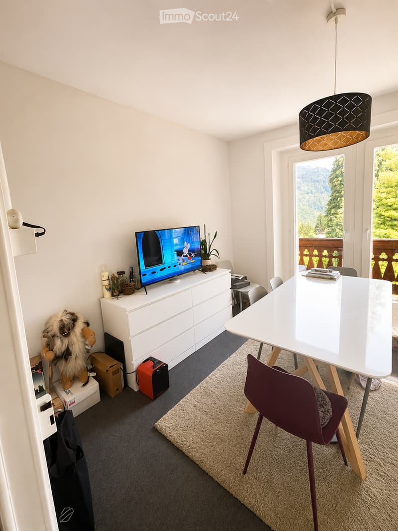 A modern, well-lit room with a white cabinet, television, dining table, chairs, and a plush toy on a stool. The room features a large window with a scenic view and a hanging lamp.