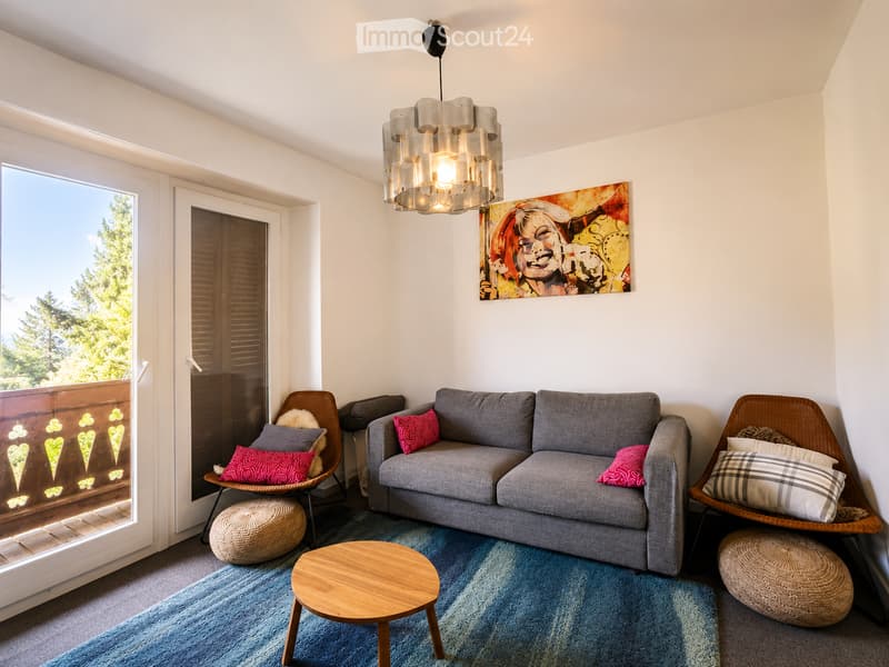 Modern living room with a grey sofa, wooden coffee table, two wicker chairs, and colorful cushions. Large windows offer a view of greenery, and a colorful abstract painting adorns the wall.
