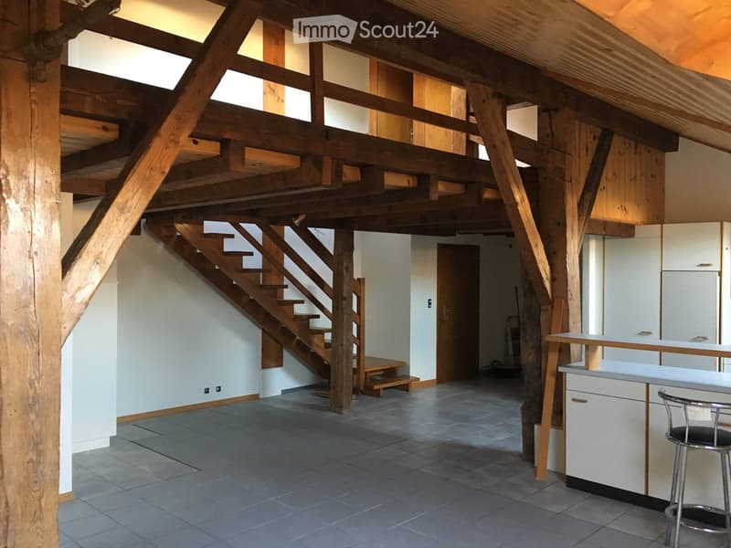 Open-plan space with exposed wooden beams, a staircase leading to an upper level, a small bench, and a kitchen area with a counter and stools.