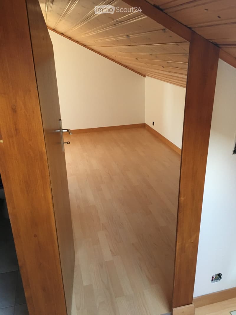 Empty attic space with wooden flooring, exposed wooden beams, and white walls.