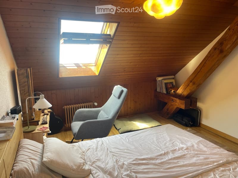 Cozy attic bedroom with wooden walls and slanted ceilings, featuring a bed, a modern armchair, a table lamp, and a floor lamp, with a skylight providing natural light.