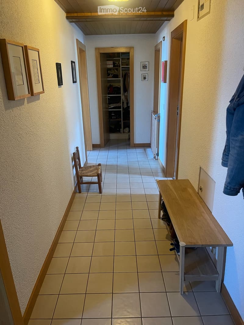 Hallway with tiled floor, wooden doors, a small wooden bench, and a wooden chair. Closet with clothes visible inside. Several framed pictures on the walls.
