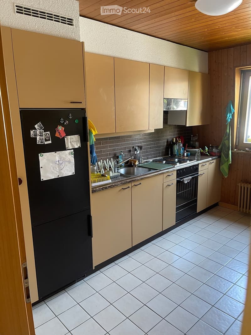 The kitchen features light-colored cabinets, a black refrigerator with magnets and notes, a sink with a faucet, and a stove with a pot. The floor is tiled, and there are various kitchen items on the counter.