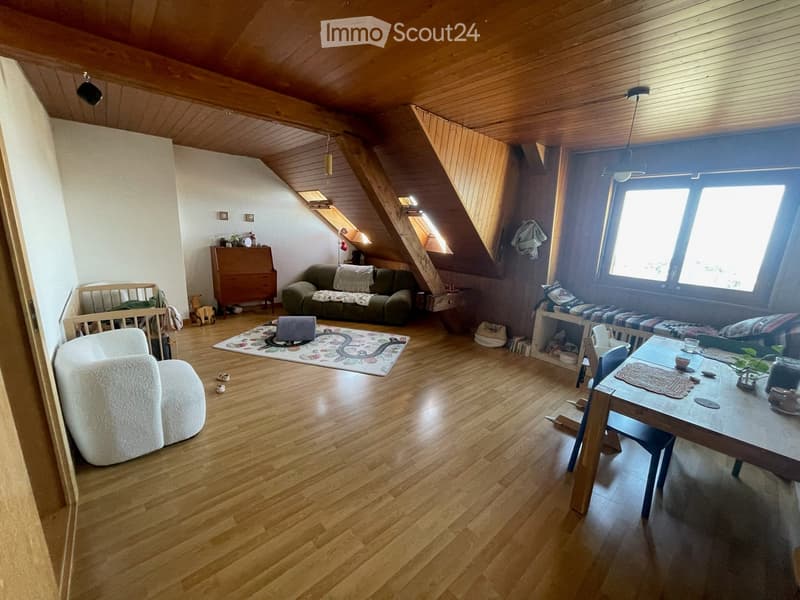 Bright, multi-functional room with wooden floors, slanted wooden ceiling, large window, sofa, dining table, baby crib, and storage cabinet.