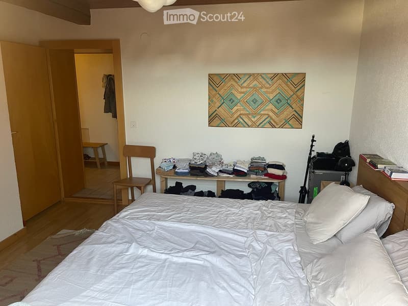 Cozy bedroom with a neatly made bed, a wooden chair, a small table with folded clothes, a lamp, and a colorful geometric painting on the wall.