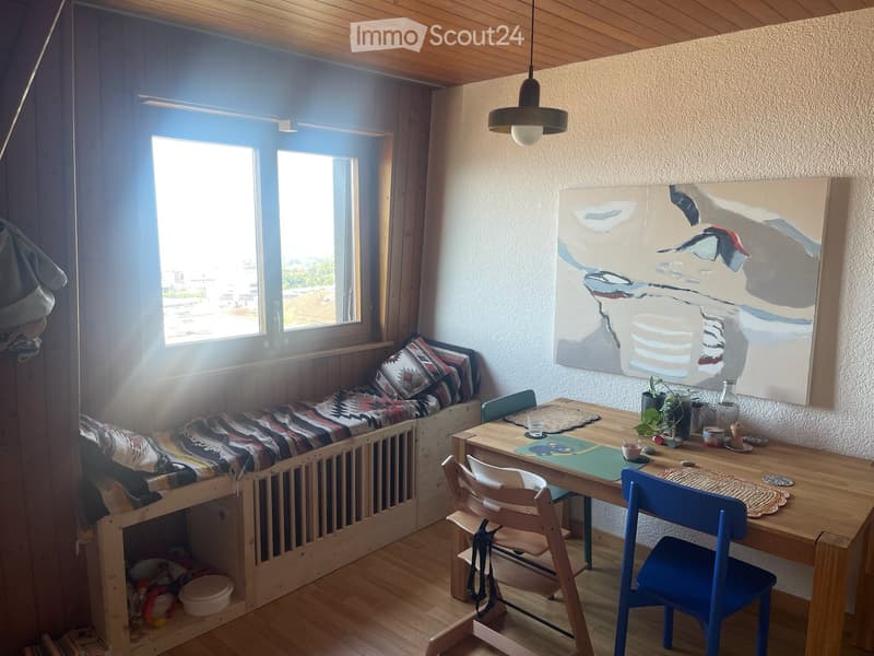 Cozy room with a daybed, small table, chairs, and artwork on the wall. Natural light from large windows.
