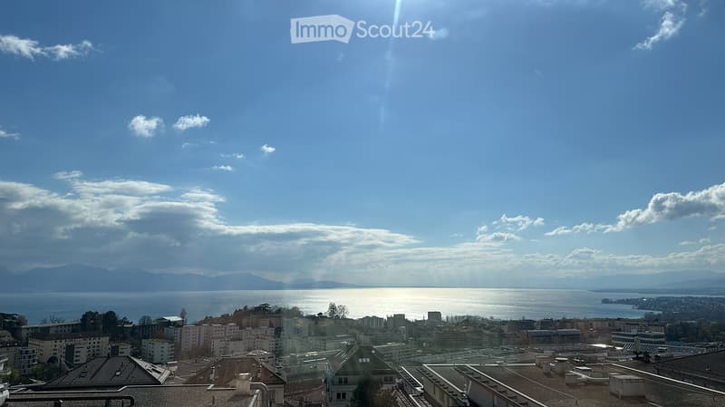 Panoramic view from a rooftop terrace overlooking a cityscape, lake, and distant mountains under a clear blue sky.