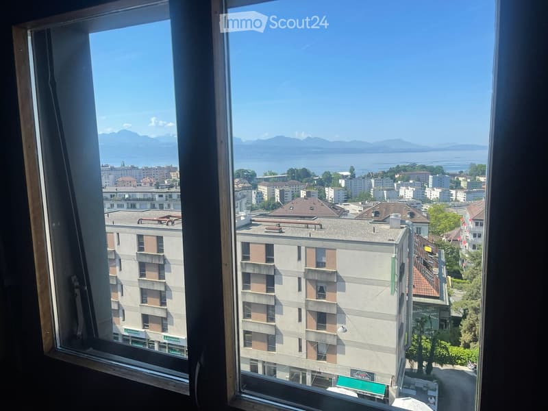 View from a window showing a cityscape with buildings, a lake, and distant mountains under a clear blue sky.