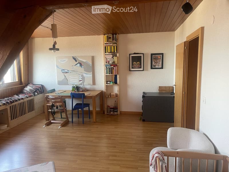 Cozy attic room with wooden floors, slanted ceilings, and large windows. Features a built-in bench seating area, a wooden dining table with chairs, a bookshelf, and a small cabinet.